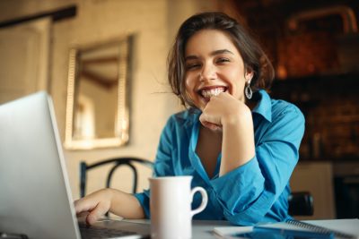 cheerful attractive young woman enjoying distant work sitting desk using portable computer drinking coffee pretty female blogger working from home uploading video her channel smiling